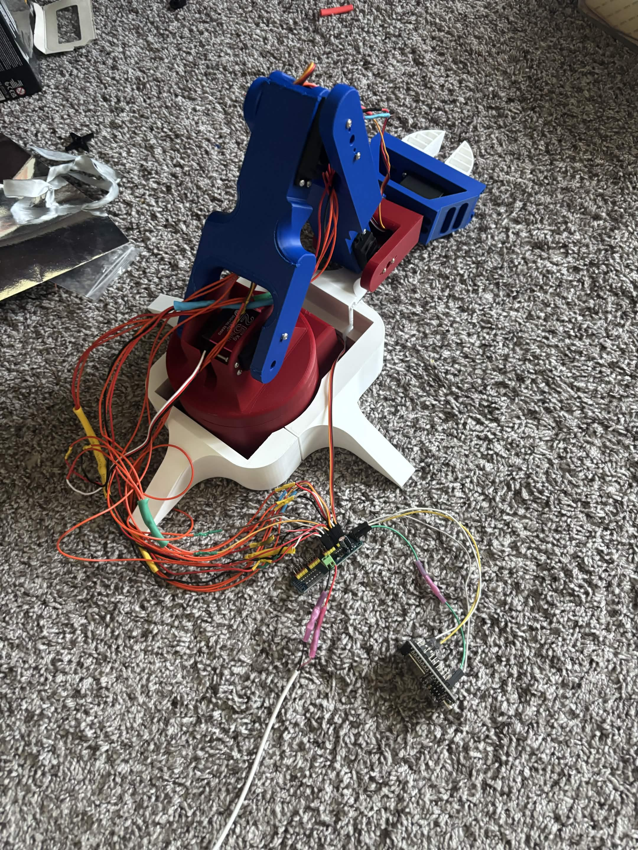 Robotic arm mounted on a broad white stabilizing base with its servo electronics and controller wired beside it